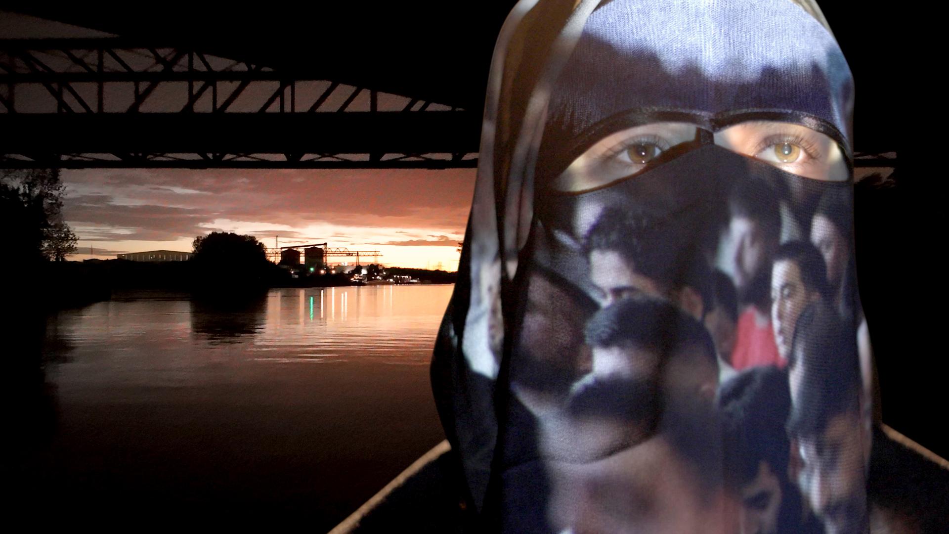 Eine Frau mit Burka, auf der Bilder projiziert werden, vor einer Flusslandschaft 
