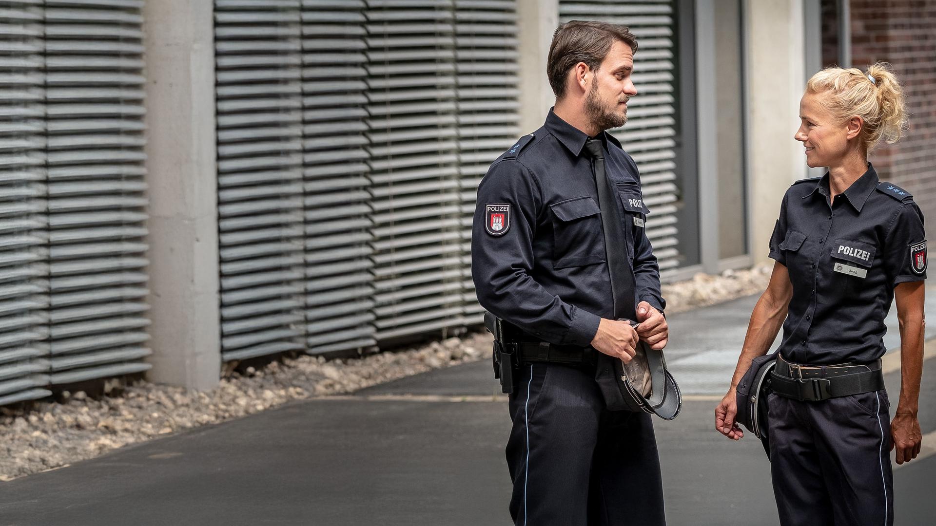"Notruf Hafenkante - Blutsbrüder": Kris Freiberg (Marc Barthel) und Franzi Jung (Rhea Harder-Vennewald) stehen nebeneinander und grinsen sich an. Im Hintergrund ist ein Bürgersteig und Häuserfronten zu sehen.