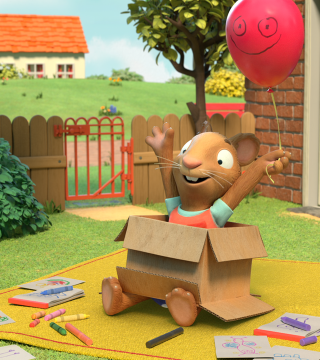 Posy sitzt freudestrahlend in einem Pappkarton und hält einen roten Luftballon an einer Schnur, der ein grinsendes Gesicht hat.