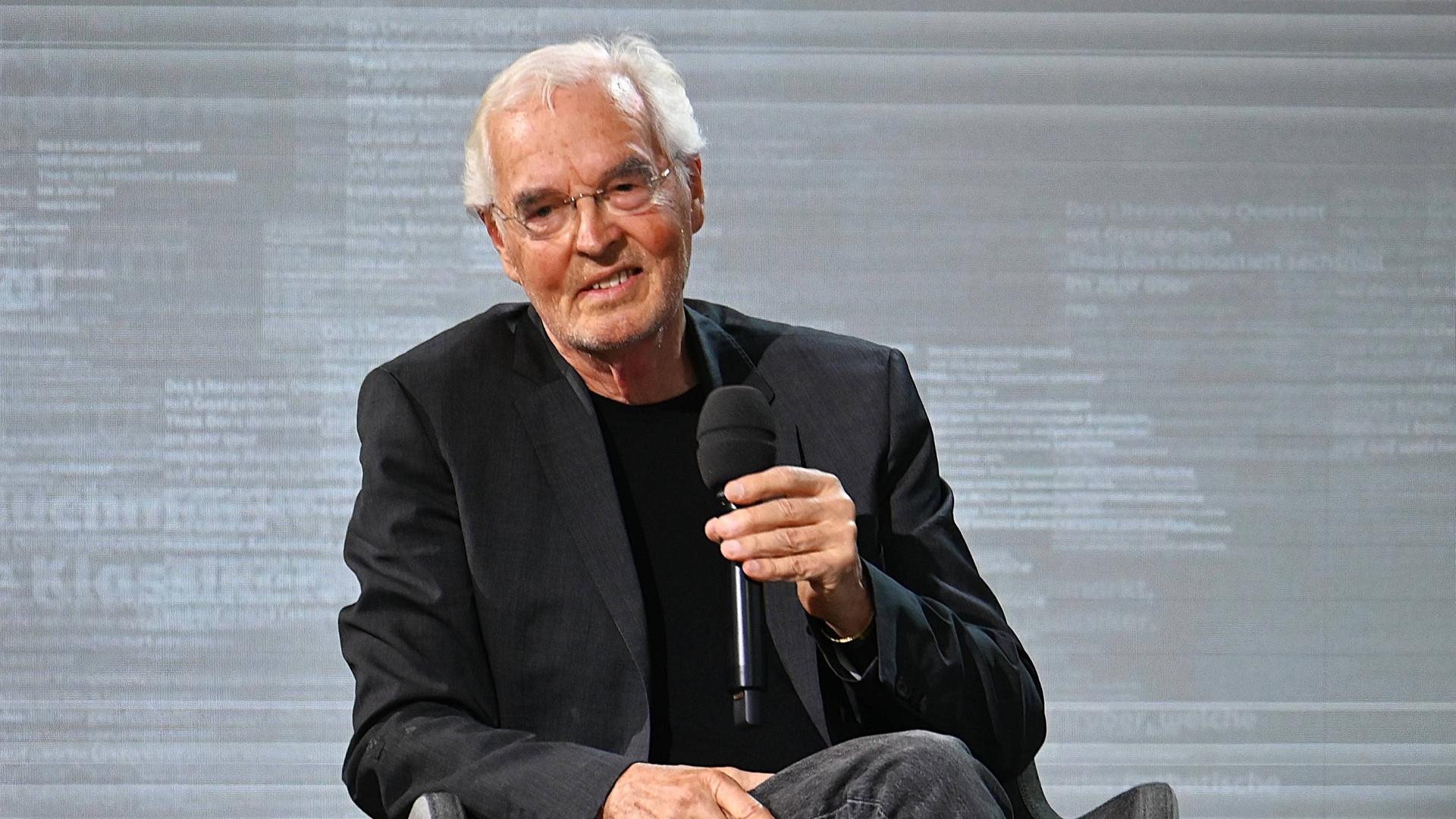 Ein älterer Mann mit weißen Haaren und im dunklen Anzug sitzt auf einem Stuhl und hält ein Mikrofon in der Hand.