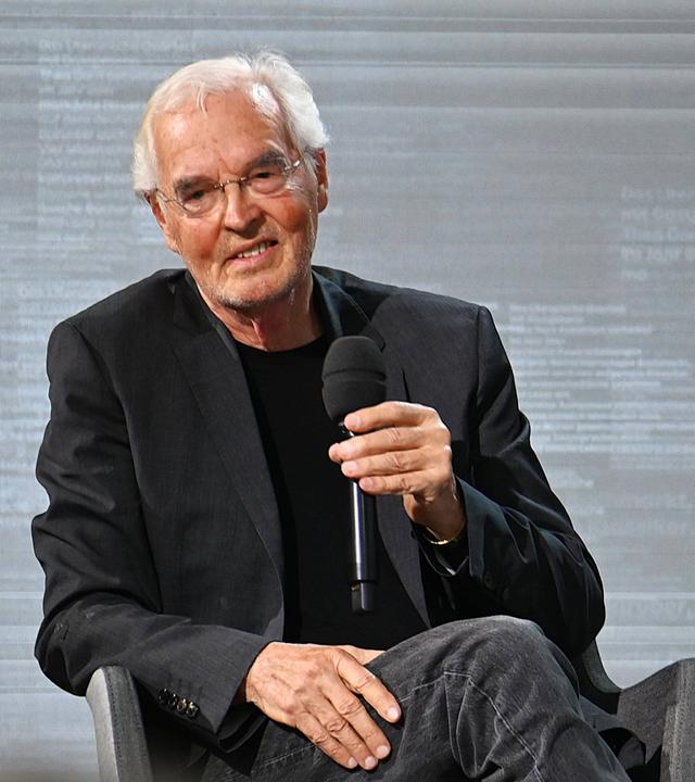 Ein älterer Mann mit weißen Haaren und im dunklen Anzug sitzt auf einem Stuhl und hält ein Mikrofon in der Hand.