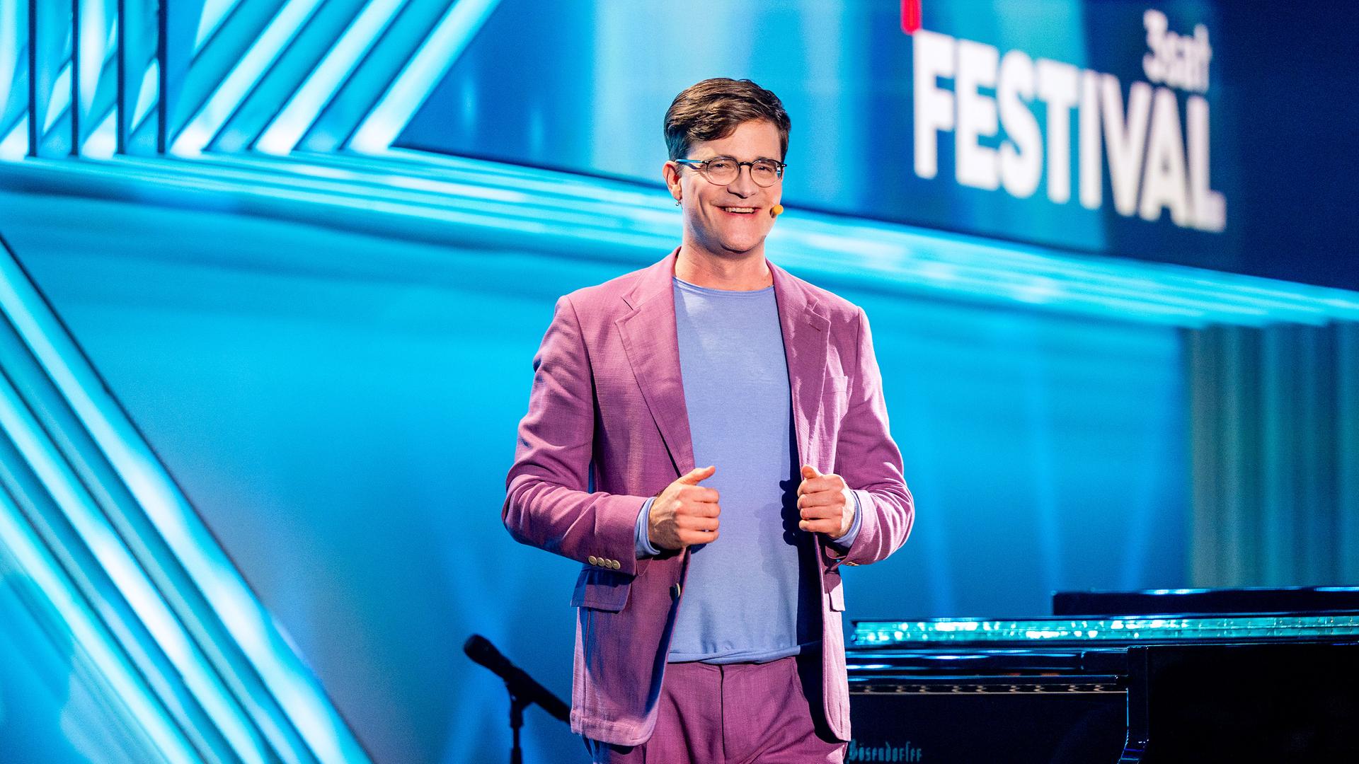 Kabarettist steht mittig auf der Bühne, er lächelt und blickt in Richtung der Kamera. Sein Anzug ist altrosa und er trägt ein hellblaues Shirt. Der Hintergrund ist blau beleuchtet.