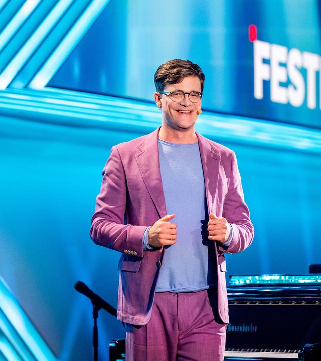 Kabarettist steht mittig auf der Bühne, er lächelt und blickt in Richtung der Kamera. Sein Anzug ist altrosa und er trägt ein hellblaues Shirt. Der Hintergrund ist blau beleuchtet.