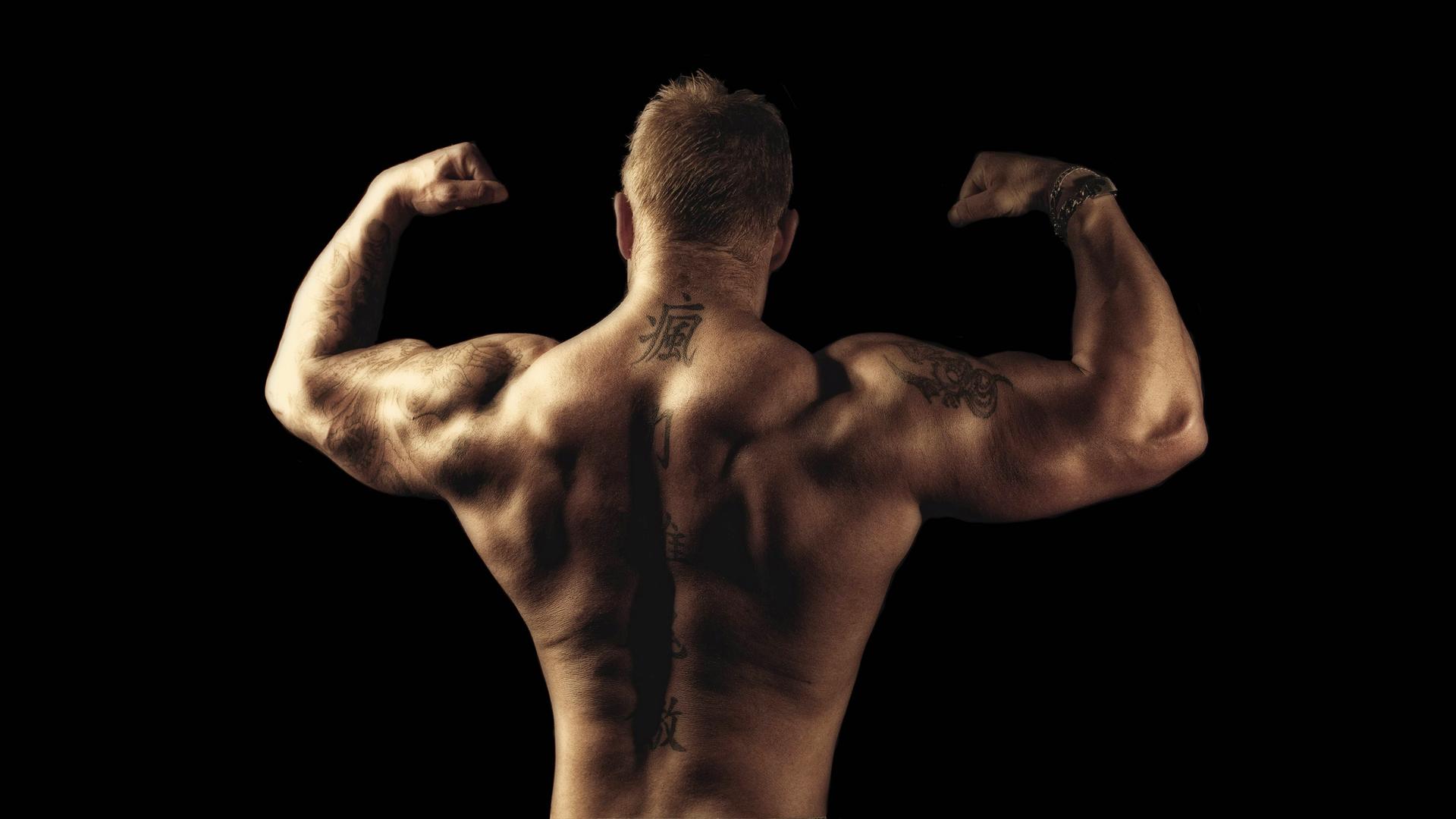 Rückansicht des Oberkörpers eines Bodybuilders mit Tatoos, der mit erhobenen Armen seine Muskeln spielen lässt.