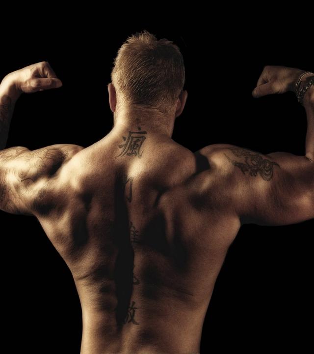 Rückansicht des Oberkörpers eines Bodybuilders mit Tatoos, der mit erhobenen Armen seine Muskeln spielen lässt.