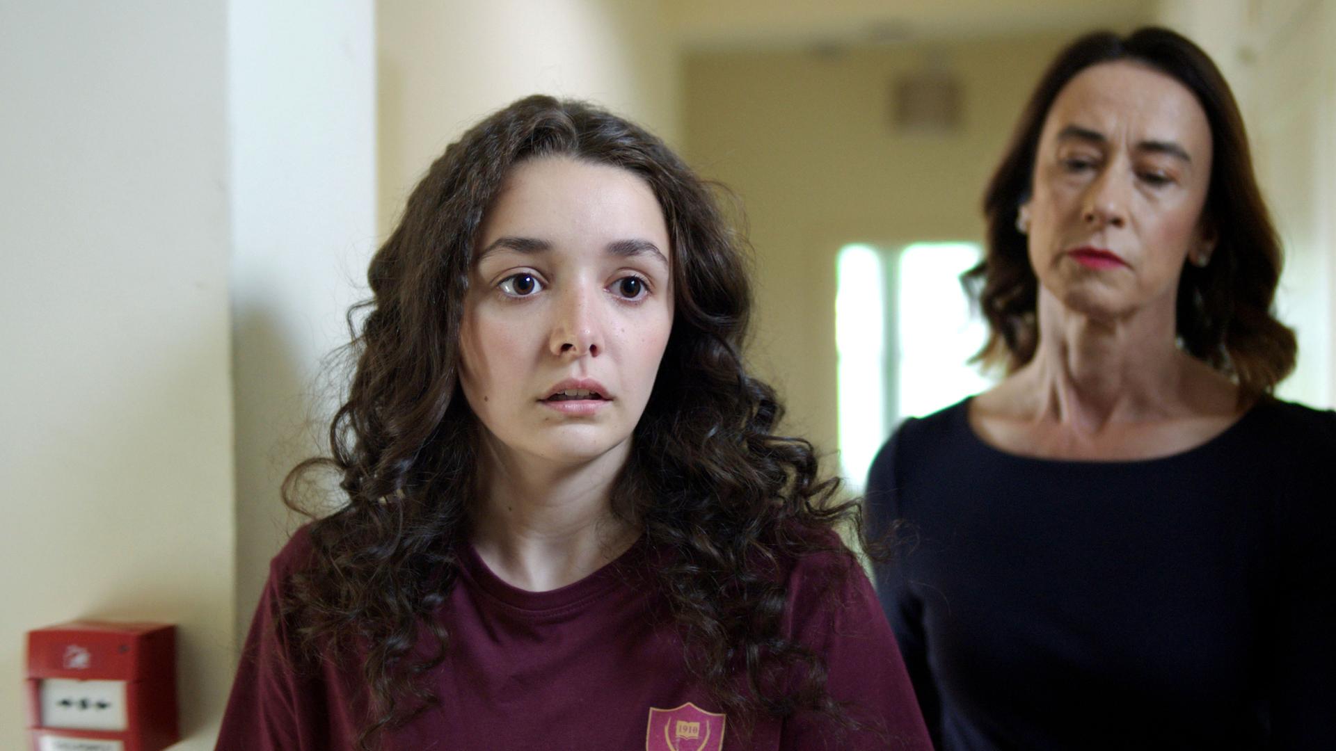 In einem weiß-gestrichenem Schulkorridor steht Addie mit fassungslosem und schockiertem Blick. Etwas versetzt hinter ihr steht die Lehrerin Miss Murphy. Sie schaut mit strengem Blick auf Addie herab.