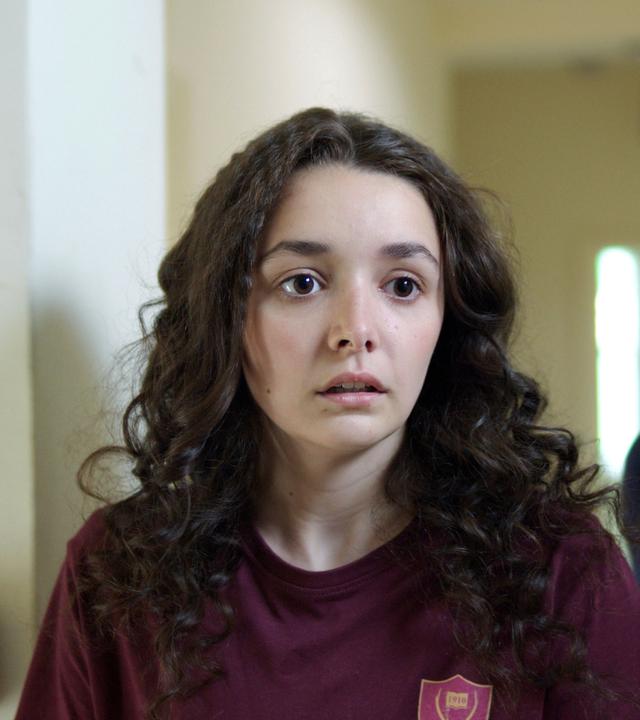 In einem weiß-gestrichenem Schulkorridor steht Addie mit fassungslosem und schockiertem Blick. Etwas versetzt hinter ihr steht die Lehrerin Miss Murphy. Sie schaut mit strengem Blick auf Addie herab.