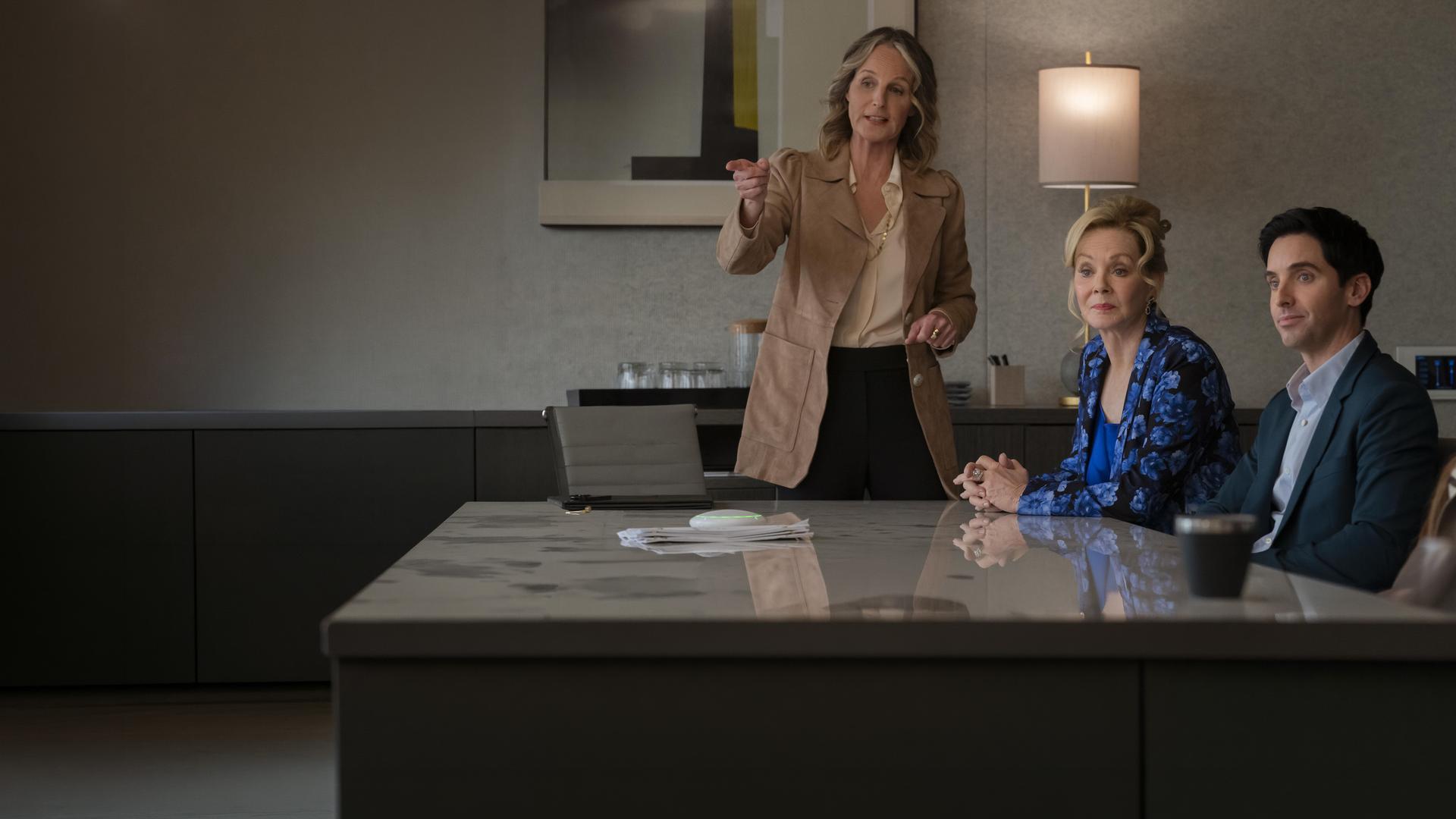 "Hacks - Bombensicher": Deborah (Jean Smart) sitzt in einer blau gemusterten Bluse neben Jimmy (Paul W. Downs) in einem Raum. Neben ihr steht Winnie Landell (Helen Hunt) die gestikulierend spricht.