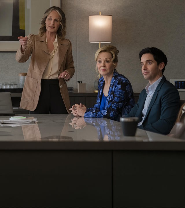 "Hacks - Bombensicher": Deborah (Jean Smart) sitzt in einer blau gemusterten Bluse neben Jimmy (Paul W. Downs) in einem Raum. Neben ihr steht Winnie Landell (Helen Hunt) die gestikulierend spricht.