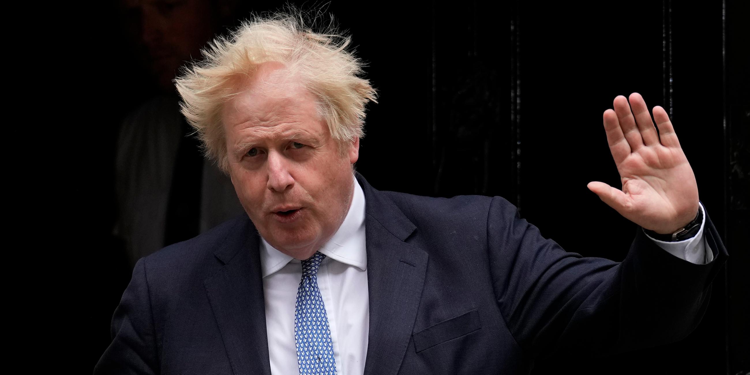 Boris Johnson mit zerzausten Haaren hebt die Linke Hand und hat zwei Ordner unter dem rechten Arm