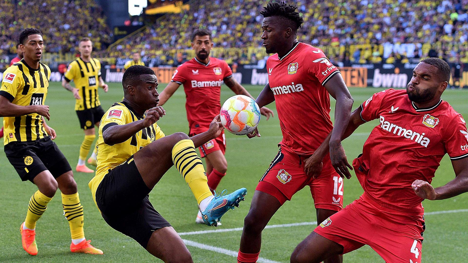 Youssoufa Moukoko (l, Dortmund) in Aktion gegen Jonathan Tah (r, Leverkusen) und Edmond Tapsoba (m, Leverkusen) am 06.08.2022.