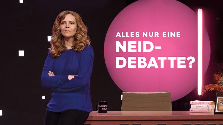 Sarah Bosetti steht auf der linken Seite des Bildes. Sie trägt ein dunkelblaues Oberteil und lehnt mit der Hüfte an einem Schreibtisch. Auf der linken Seite des Tisches steht eine Tasse, rechts liegen drei Bücher übereinander. 