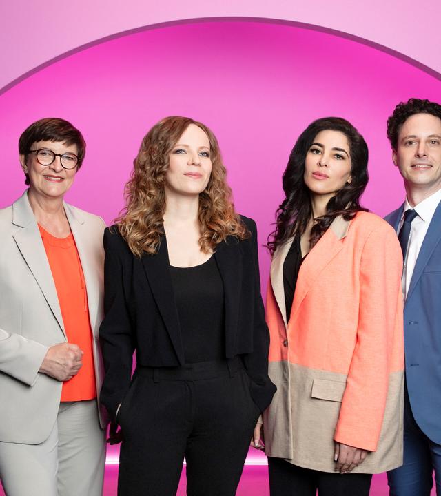Auf dem Bild ist Sarah Bosetti als zweite von links zu sehen. Der Hintergrund ist rosa und in der Mitte blau. Links neben Sarah Bosetti steht Saskia Esken. Rechts von Sarah steht Samira El Ouassil und neben ihr ist Matthias Renger zu sehen.