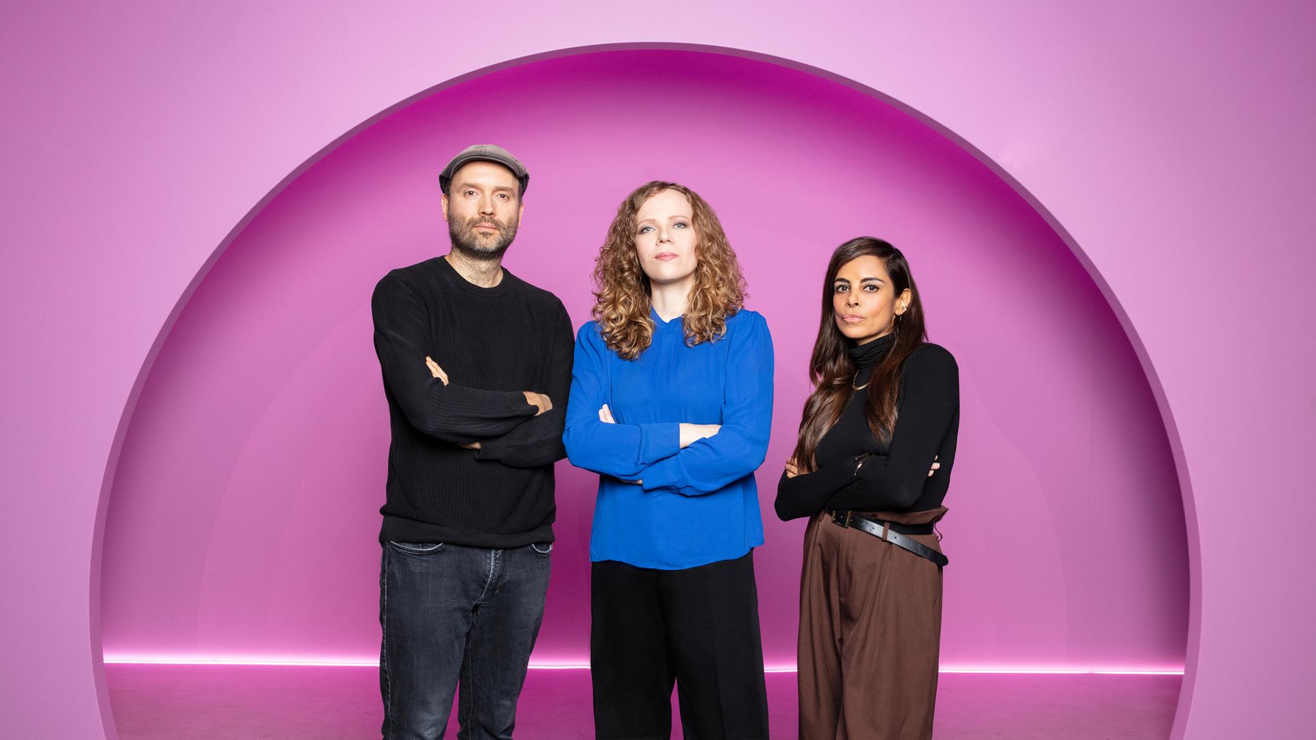 Sarah Bosetti steht in der Mitte des Bildes. Auf ihrer linken Seite steht Marc-Uwe Kling. Rechts von ihr ist Collien Fernandes. Der Hintergrund ist rosa.