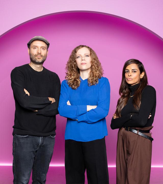 Sarah Bosetti steht in der Mitte des Bildes. Auf ihrer linken Seite steht Marc-Uwe Kling. Rechts von ihr ist Collien Fernandes. Der Hintergrund ist rosa.