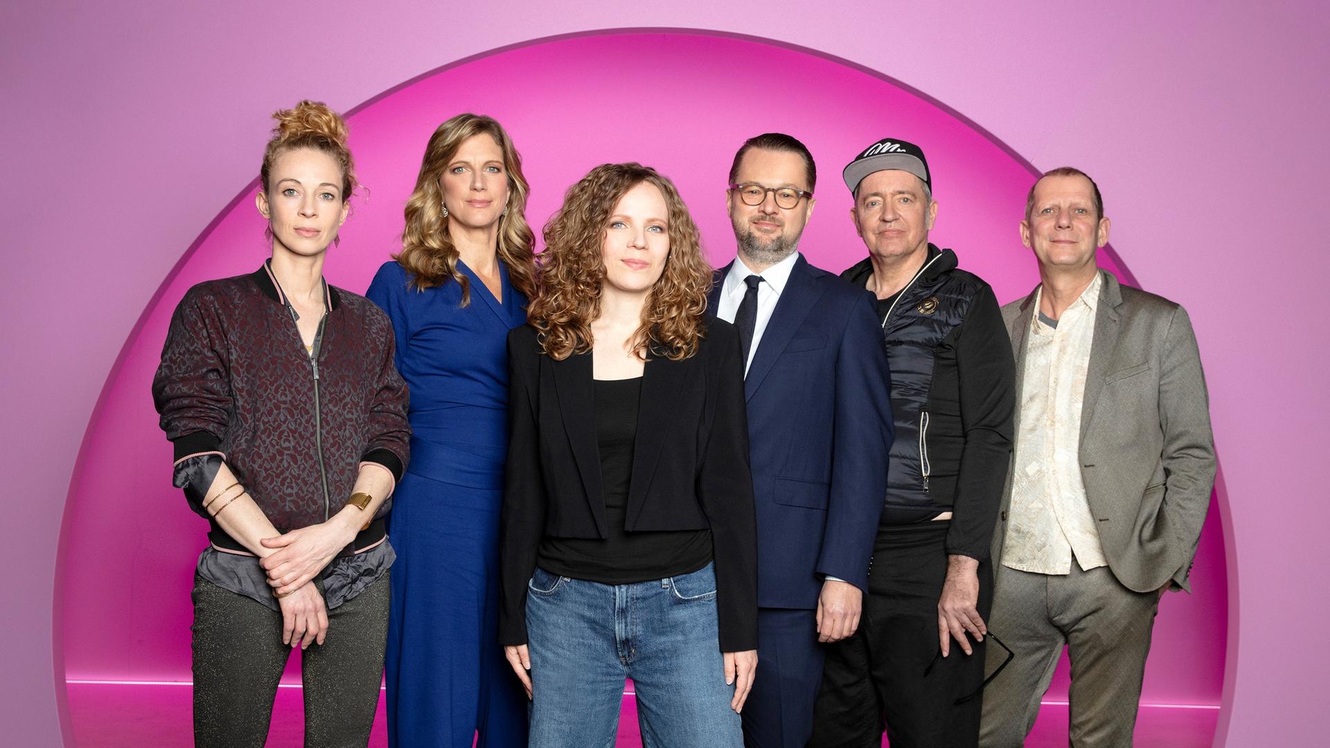 Sarah Bosetti steht in der Mitte des Bildes. Der Hintergrund ist rosa. Es sind 6 Personen abgebildet, zwei links von Sarah Bosetti und drei auf ihrer rechten Seite. Sarah Bosetti trägt eine Jeans, ein schwarzes Oberteil sowie einen kurzen schwarzen Blazer.