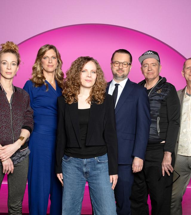 Sarah Bosetti steht in der Mitte des Bildes. Der Hintergrund ist rosa. Es sind 6 Personen abgebildet, zwei links von Sarah Bosetti und drei auf ihrer rechten Seite. Sarah Bosetti trägt eine Jeans, ein schwarzes Oberteil sowie einen kurzen schwarzen Blazer.