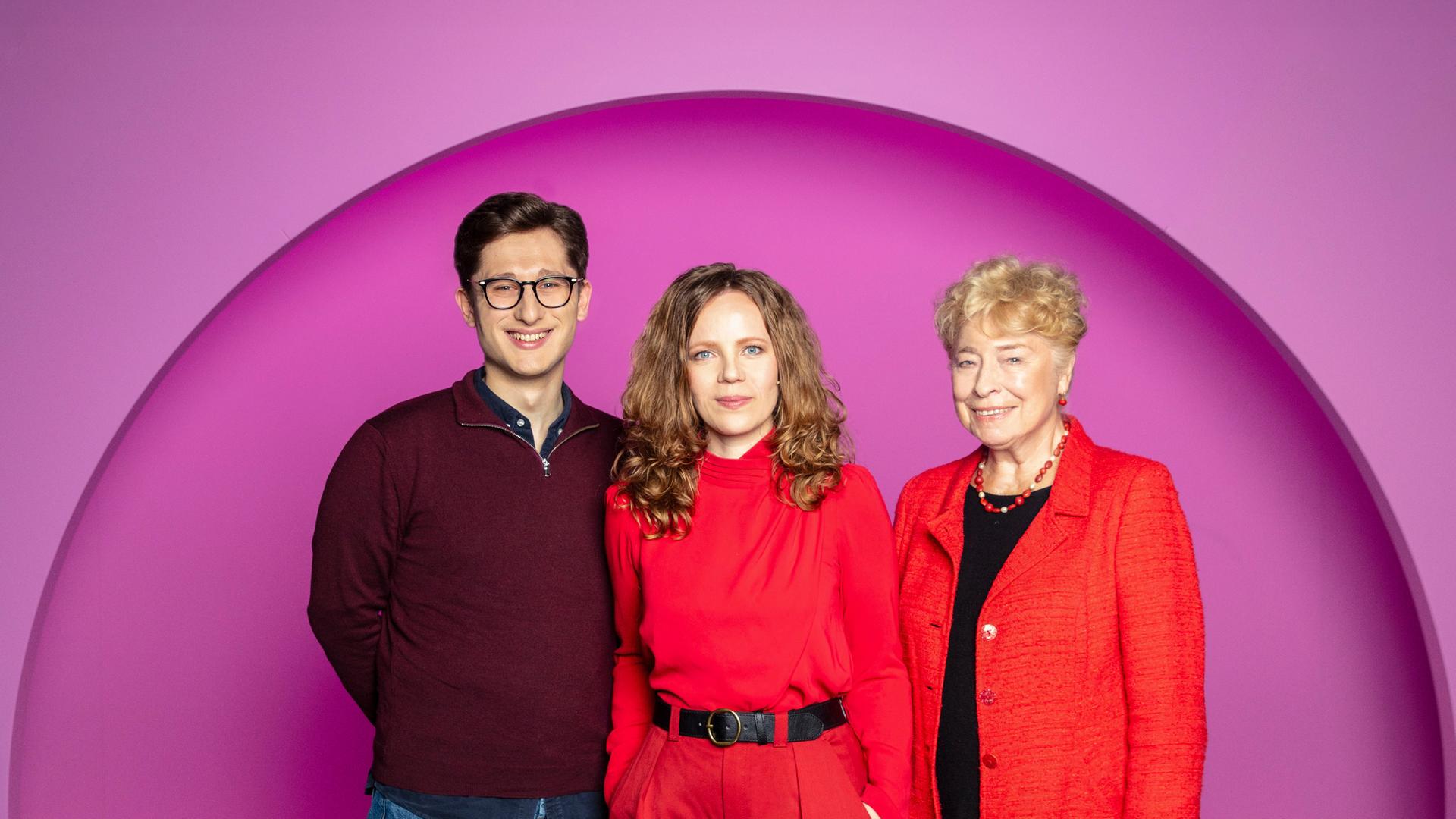 Sarah Bosetti steht in der Mitte des Bildes. Der Hintergrund ist rosa. Es sind drei Personen abgebildet. Links steht Philipp Türmer und rechts Gesine Schwan.