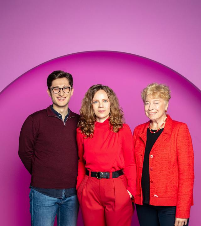 Sarah Bosetti steht in der Mitte des Bildes. Der Hintergrund ist rosa. Es sind drei Personen abgebildet. Links steht Philipp Türmer und rechts Gesine Schwan.