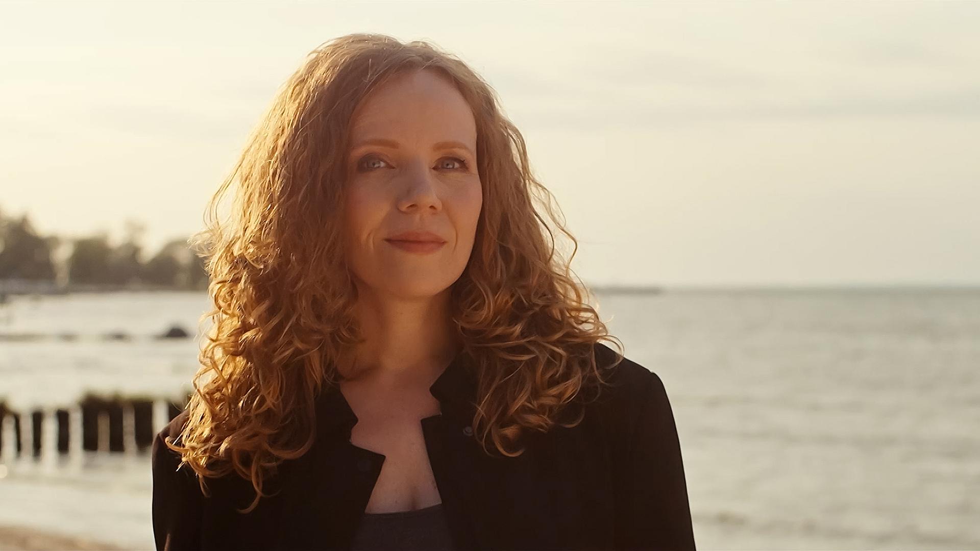 Sarah Bosetti steht in der Mitte des Bildes. Im Hintergrund ist ein Sonnenuntergang und Wasser, das Licht ist hell. Sarah Bosetti trägt eine schwarze Jacke, ihre Haare sind offen. 