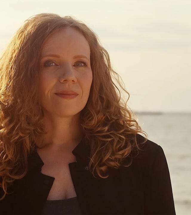 Sarah Bosetti steht in der Mitte des Bildes. Im Hintergrund ist ein Sonnenuntergang und Wasser, das Licht ist hell. Sarah Bosetti trägt eine schwarze Jacke, ihre Haare sind offen. 