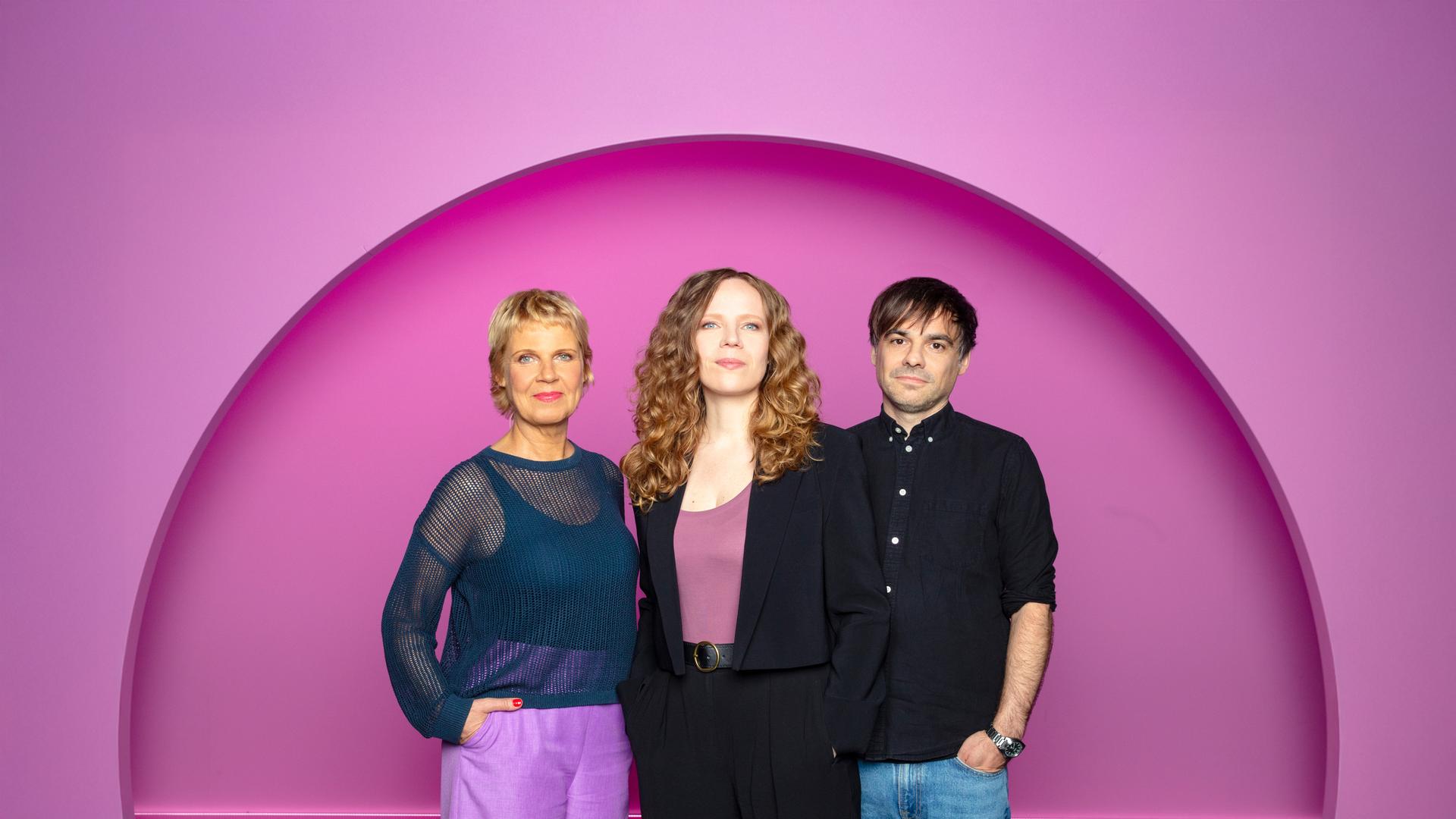 Sarah Bosetti steht in der Mitte des Bildes. Links von ihr steht Cordula Stratmann und rechts von Sarah Bosetti ist Sebastian Lehmann. Der Hintergrund ist rosa.