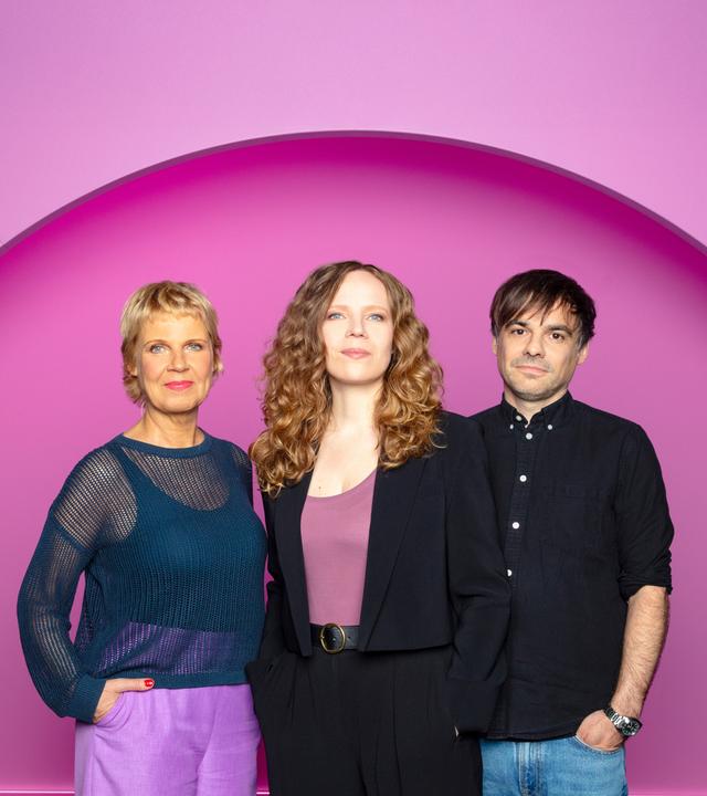 Sarah Bosetti steht in der Mitte des Bildes. Links von ihr steht Cordula Stratmann und rechts von Sarah Bosetti ist Sebastian Lehmann. Der Hintergrund ist rosa.