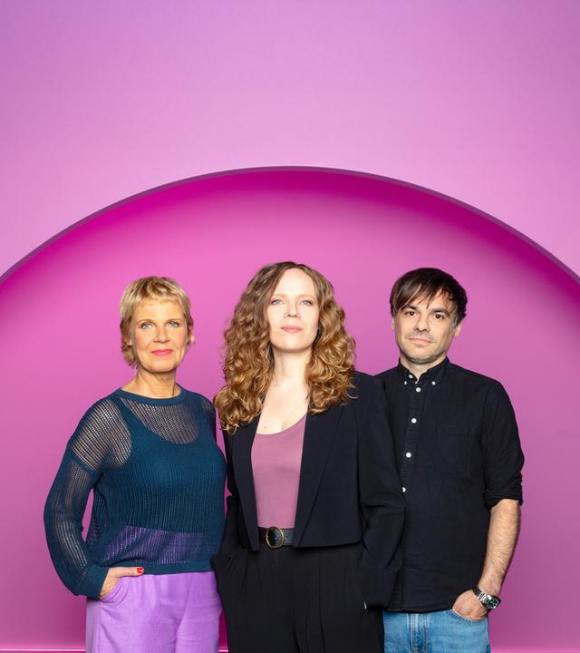 Sarah Bosetti steht in der Mitte des Bildes. Links von ihr steht Cordula Stratmann und rechts von Sarah Bosetti ist Sebastian Lehmann. Der Hintergrund ist rosa.