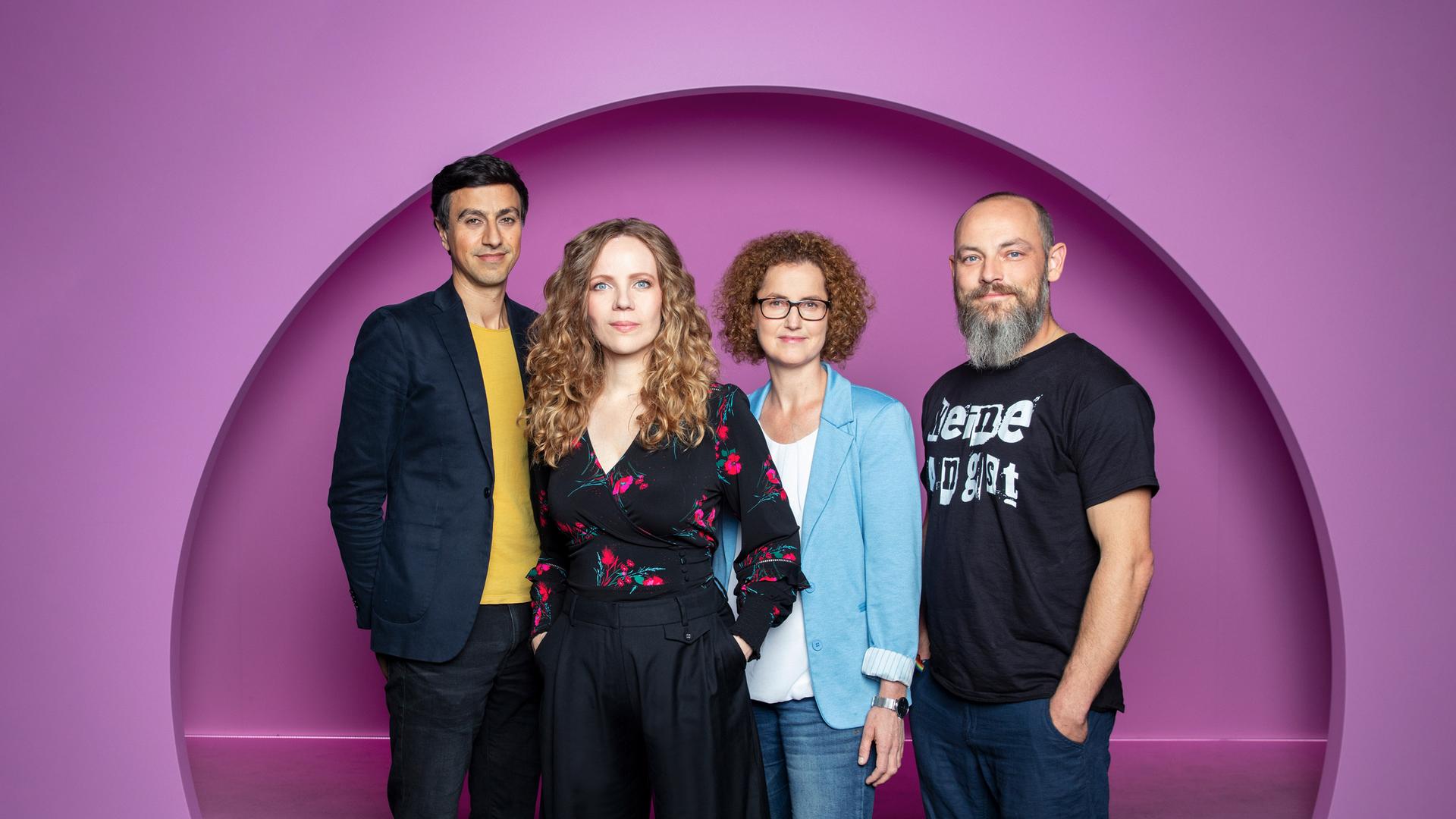 Auf dem Bild stehen vier Personen nebeneinander. Sarah Bosetti ist die zweite Person von Link.  Sie trägt ihr Haar offen und ist schwarz gekleidet. Der Hintergrund ist rosa.