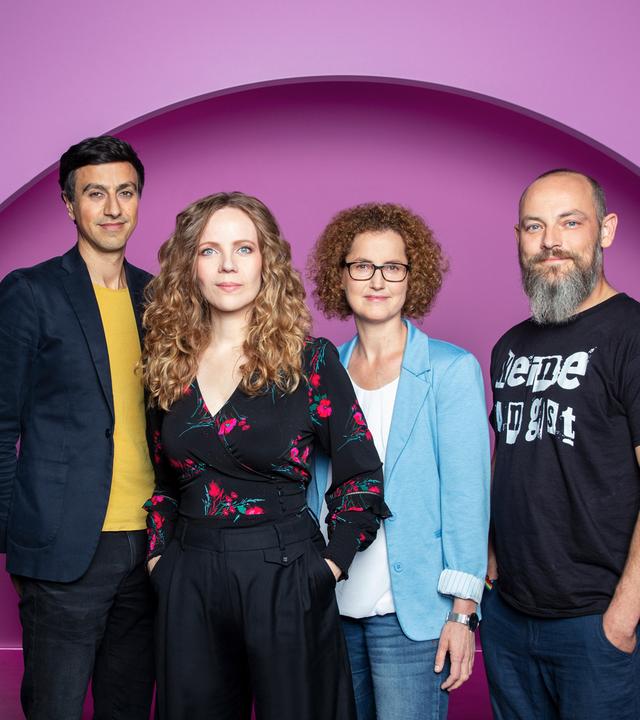 Auf dem Bild stehen vier Personen nebeneinander. Sarah Bosetti ist die zweite Person von Link.  Sie trägt ihr Haar offen und ist schwarz gekleidet. Der Hintergrund ist rosa.
