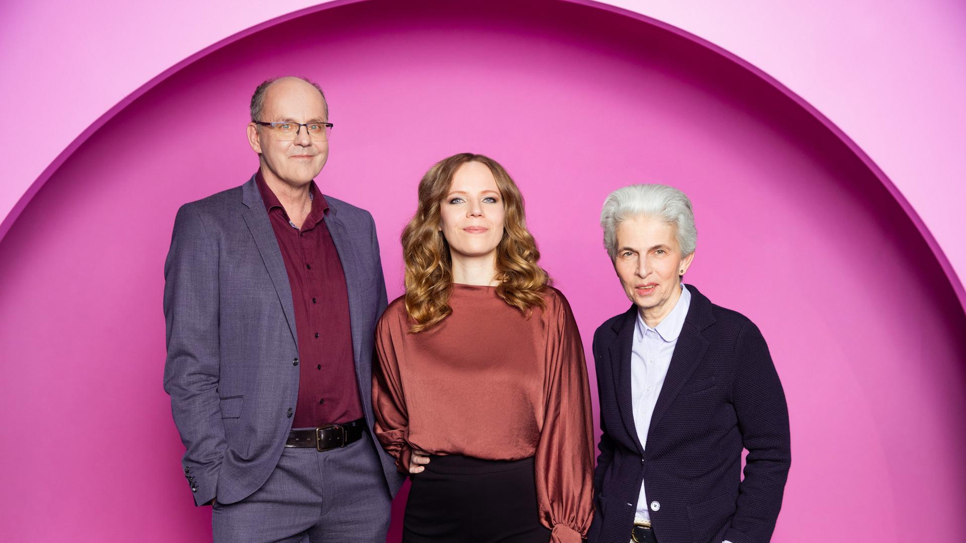 Auf dem Bild ist Sarah Bosetti mittig zu sehen. Links neben ihr steht Andreas Hasenclever. Auf der rechten Seite des Bildes ist Marie-Agnes Strack-Zimmermann. Der Hintergrund ist rosa.