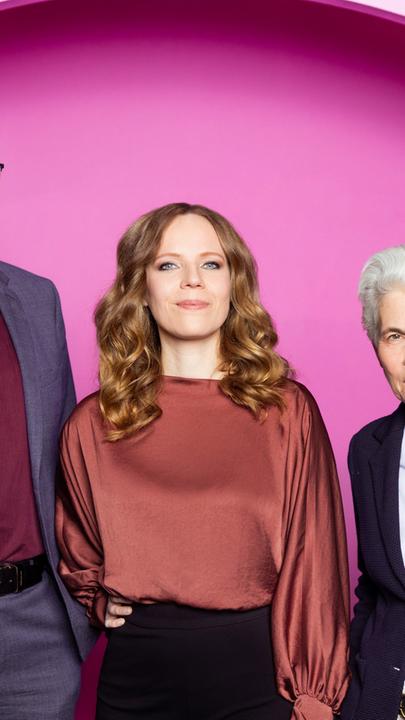 Auf dem Bild ist Sarah Bosetti mittig zu sehen. Links neben ihr steht Andreas Hasenclever. Auf der rechten Seite des Bildes ist Marie-Agnes Strack-Zimmermann. Der Hintergrund ist rosa.