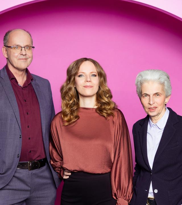 Auf dem Bild ist Sarah Bosetti mittig zu sehen. Links neben ihr steht Andreas Hasenclever. Auf der rechten Seite des Bildes ist Marie-Agnes Strack-Zimmermann. Der Hintergrund ist rosa.