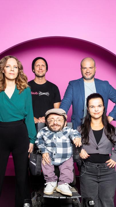 Auf dem Bild ist Sarah Bosetti auf der linken Seite zu sehen. Neben ihr stehen vier weitere Personen. Der Hintergrund ist rosa. 