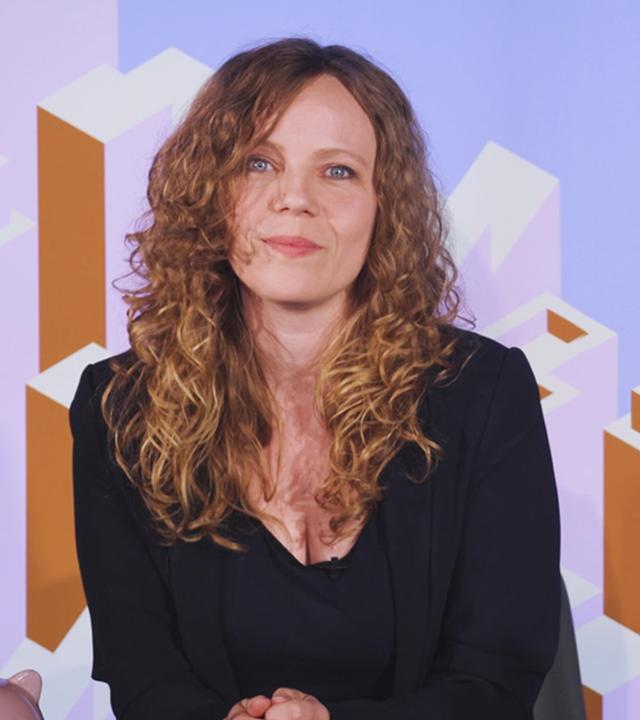 Sarah Bosetti sitzt an einem Schreibtisch. Rechts neben ihr ist Friedrich Merz in einer Blase abgebildet.