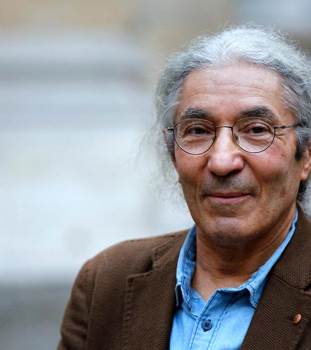 Porträtfoto eines Mannes mit zurückgebundenen grauen Haaren und runder Metallbrille, der in die Kamera lächelt.