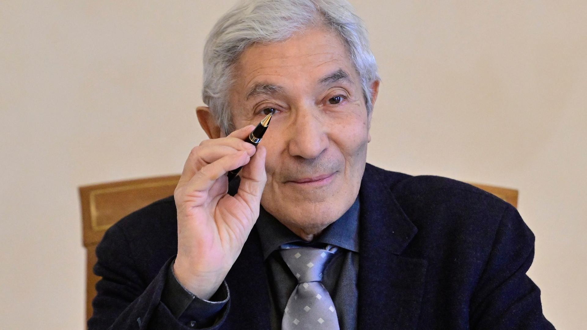 Porträtfoto eines Mannes mit zurückgebundenen grauen Haaren und runder Metallbrille, der in die Kamera lächelt.