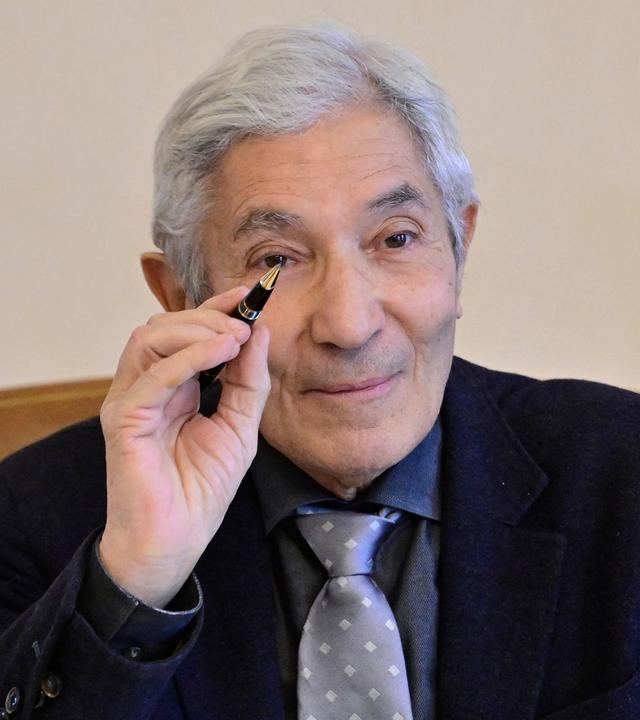 Porträtfoto eines Mannes mit zurückgebundenen grauen Haaren und runder Metallbrille, der in die Kamera lächelt.