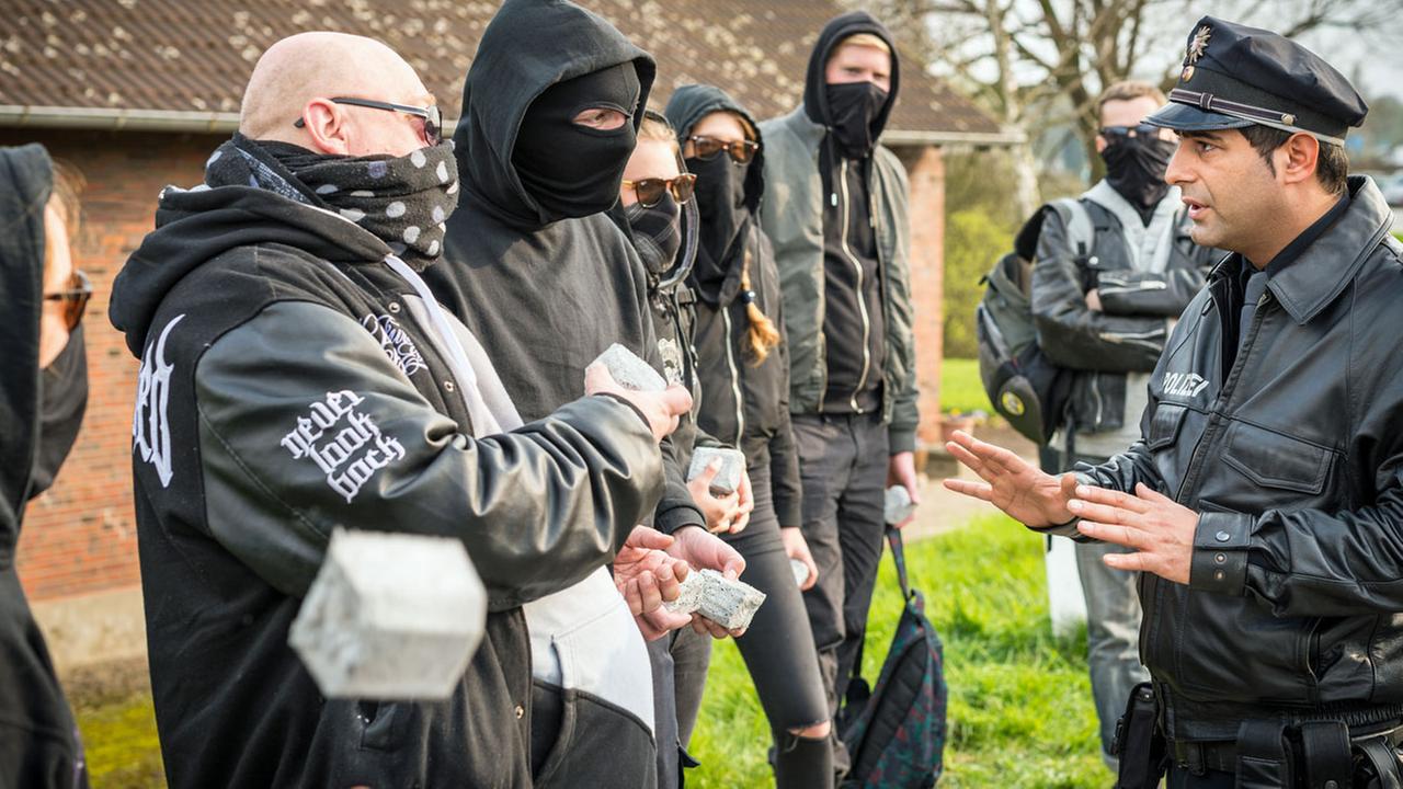 "Notruf Hafenkante - Brandstifter": Der Polizist Tarik Coban (Serhat Cokgezen) steht, die Hände besänftigend hochhaltend, einer Gruppe vermummter Hooligans gegenüber.