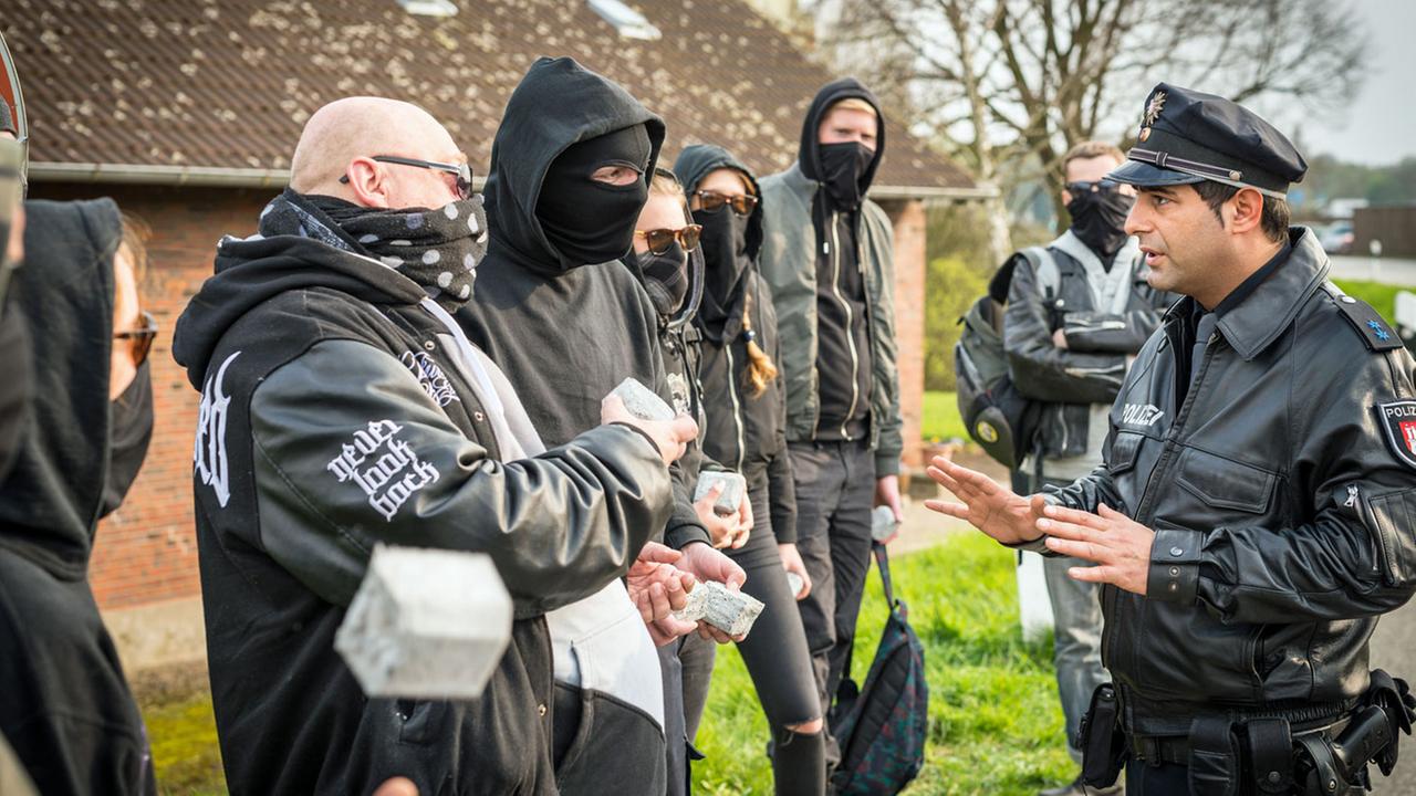 "Notruf Hafenkante - Brandstifter": Der Polizist Tarik Coban (Serhat Cokgezen) steht, die Hände besänftigend hochhaltend, einer Gruppe vermummter Hooligans gegenüber.