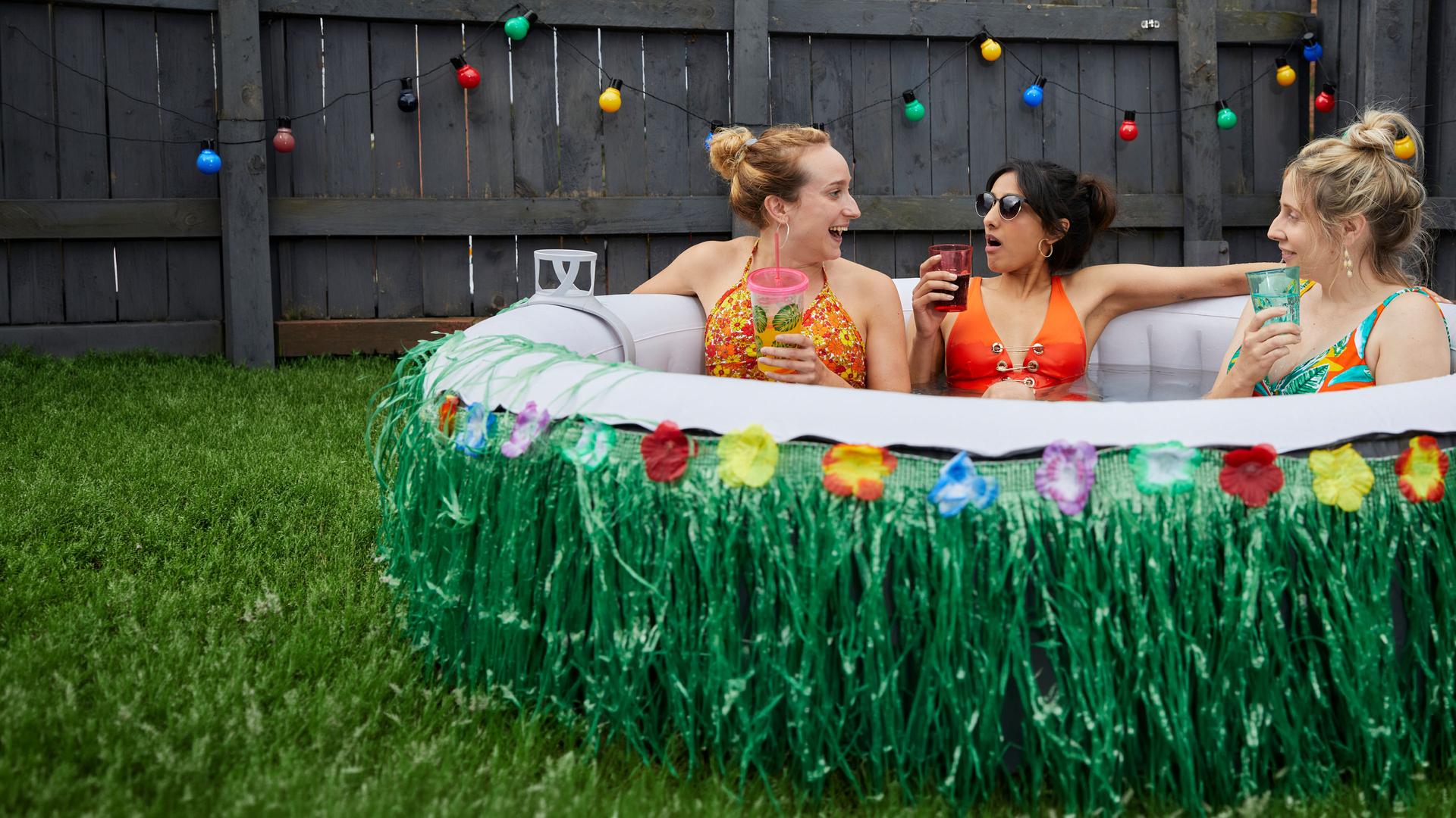 "Hullraisers - Breadcakes": Toni (Leah Brotherhead), Rana (Taj Atwal) und Paula (Sinead Matthews)  sitzen zusammen im Bikini in einem aufblasbaren Swimmingpool und schauen sich lachend an, während sie etwas trinken.