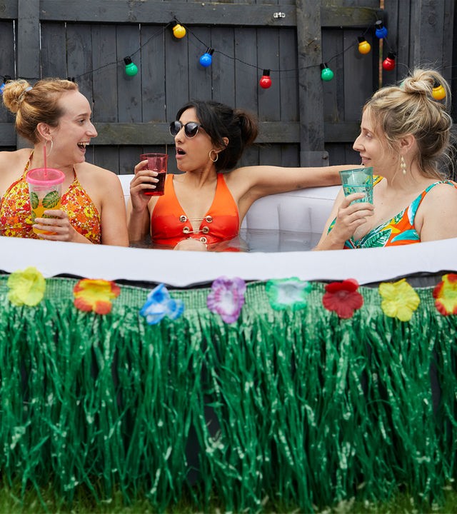 "Hullraisers - Breadcakes": Toni (Leah Brotherhead), Rana (Taj Atwal) und Paula (Sinead Matthews)  sitzen zusammen im Bikini in einem aufblasbaren Swimmingpool und schauen sich lachend an, während sie etwas trinken.