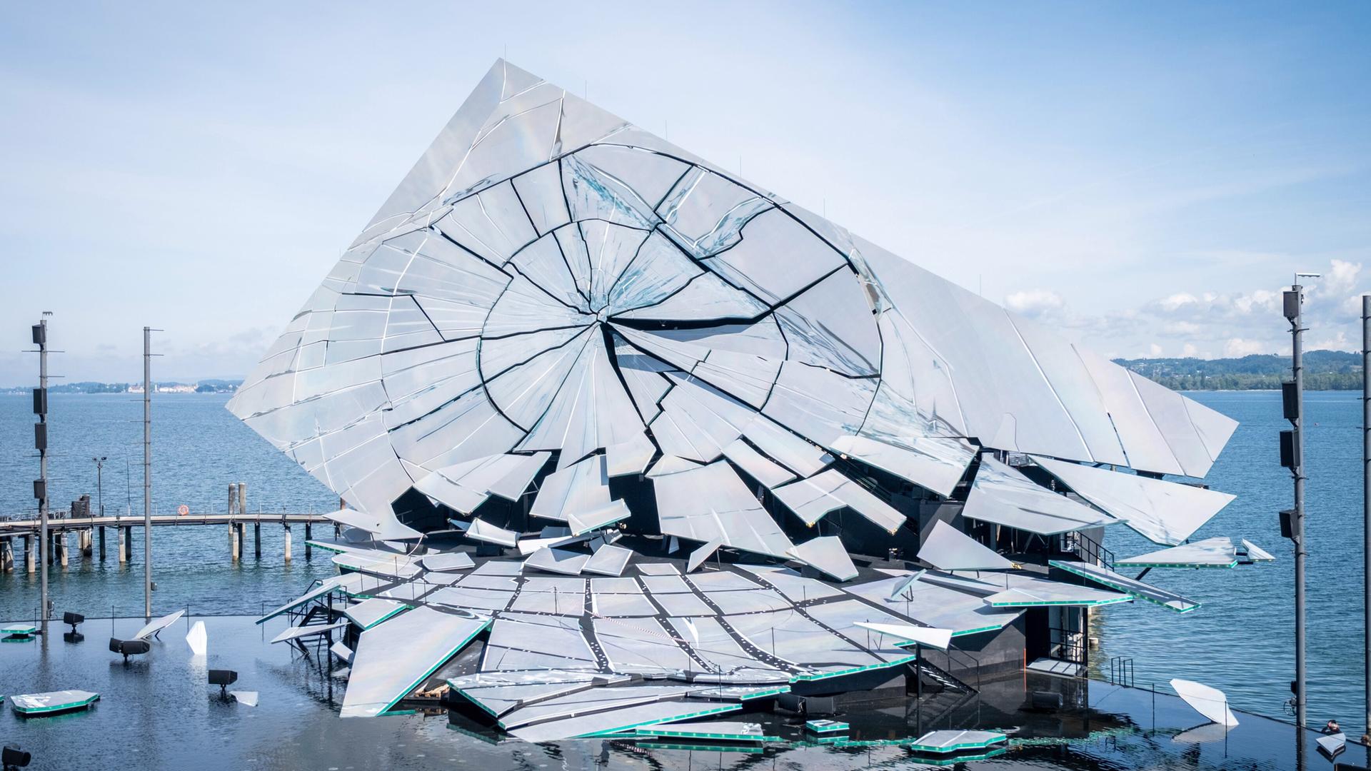 Das Bild zeigt eine schillernde, skulpturale Bühne auf dem Wasser. Die Struktur besteht aus mehreren großen, reflektierenden Flächen, die wie zerbrochene Glasstücke gestaltet sind. Sie sind asymmetrisch angeordnet und verleihen der Bühne ein dynamisches, dramatisches Aussehen. Der Hintergrund besteht aus einem klaren, blauen Himmel, unterbrochen von einigen Wolken.   Um die Bühne herum befinden sich kleinere Boote auf dem Wasser und mehrere Stützpfeiler, die möglicherweise zur Stabilisierung der Bühne dienen. Diese Pfeiler sind dunkel gehalten und heben sich vom helleren Wasser und der Bühne ab. Im Hintergrund sieht man die Uferlinie mit Bäumen und möglicherweise weiteren Gebäuden, jedoch sind diese nicht detailliert sichtbar.   Das Bild vermittelt die Atmosphäre eines feierlichen Ereignisses, da die Bühne für eine Opernaufführung konzipiert ist.