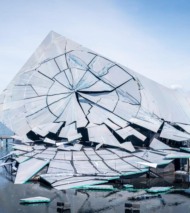 Das Bild zeigt eine schillernde, skulpturale Bühne auf dem Wasser. Die Struktur besteht aus mehreren großen, reflektierenden Flächen, die wie zerbrochene Glasstücke gestaltet sind. Sie sind asymmetrisch angeordnet und verleihen der Bühne ein dynamisches, dramatisches Aussehen. Der Hintergrund besteht aus einem klaren, blauen Himmel, unterbrochen von einigen Wolken.   Um die Bühne herum befinden sich kleinere Boote auf dem Wasser und mehrere Stützpfeiler, die möglicherweise zur Stabilisierung der Bühne dienen. Diese Pfeiler sind dunkel gehalten und heben sich vom helleren Wasser und der Bühne ab. Im Hintergrund sieht man die Uferlinie mit Bäumen und möglicherweise weiteren Gebäuden, jedoch sind diese nicht detailliert sichtbar.   Das Bild vermittelt die Atmosphäre eines feierlichen Ereignisses, da die Bühne für eine Opernaufführung konzipiert ist.