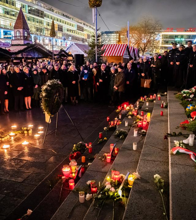 Eine Menschenmenge steht bei nacht um auf dem Boden aufgestellte Kerzen und einem Kranz vor einer Treppe, auf der noch mehr Votivkerzen stehen. Im Hintergrund ist ein Weihnachtsmarkt zu sehen.