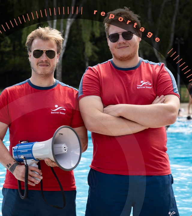 Pablo Spallek und sein Kollege Jakob stehen am Beckenrand eines Freibads. Beide tragen rote T-Shirts und blaue Shorts mit dem Logo der "Berliner Bäder" sowie eine Sonnenbrille. Pablo Spallek hält ein Megafon in der Hand. Jakob hat die Arme vor der Brust verschränkt. Im Hintergrund schwimmen Menschen im Wasser.