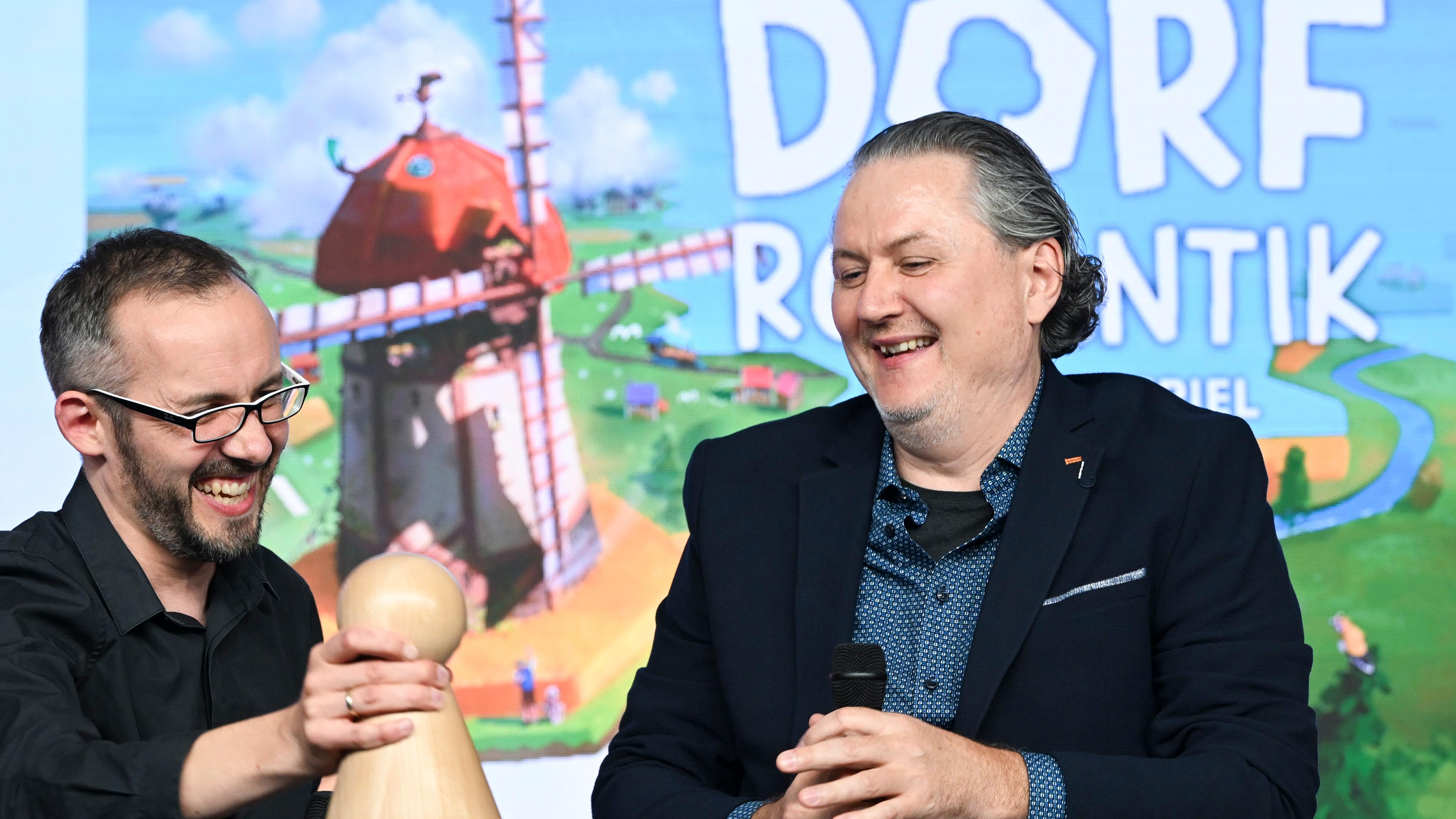 Die Spieleentwickler Lukas Zach (l) und Michael Palm vor dem Bild ihre Spiels "Dorfromantik: Das Brettspiel", das zum "Spiel des Jahres 2023" gewählt wurde