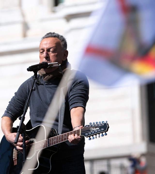 Mann spielt Gitarre und Mundharmonika, im Vordergrund Fahne mit durchgestrichener Krone.