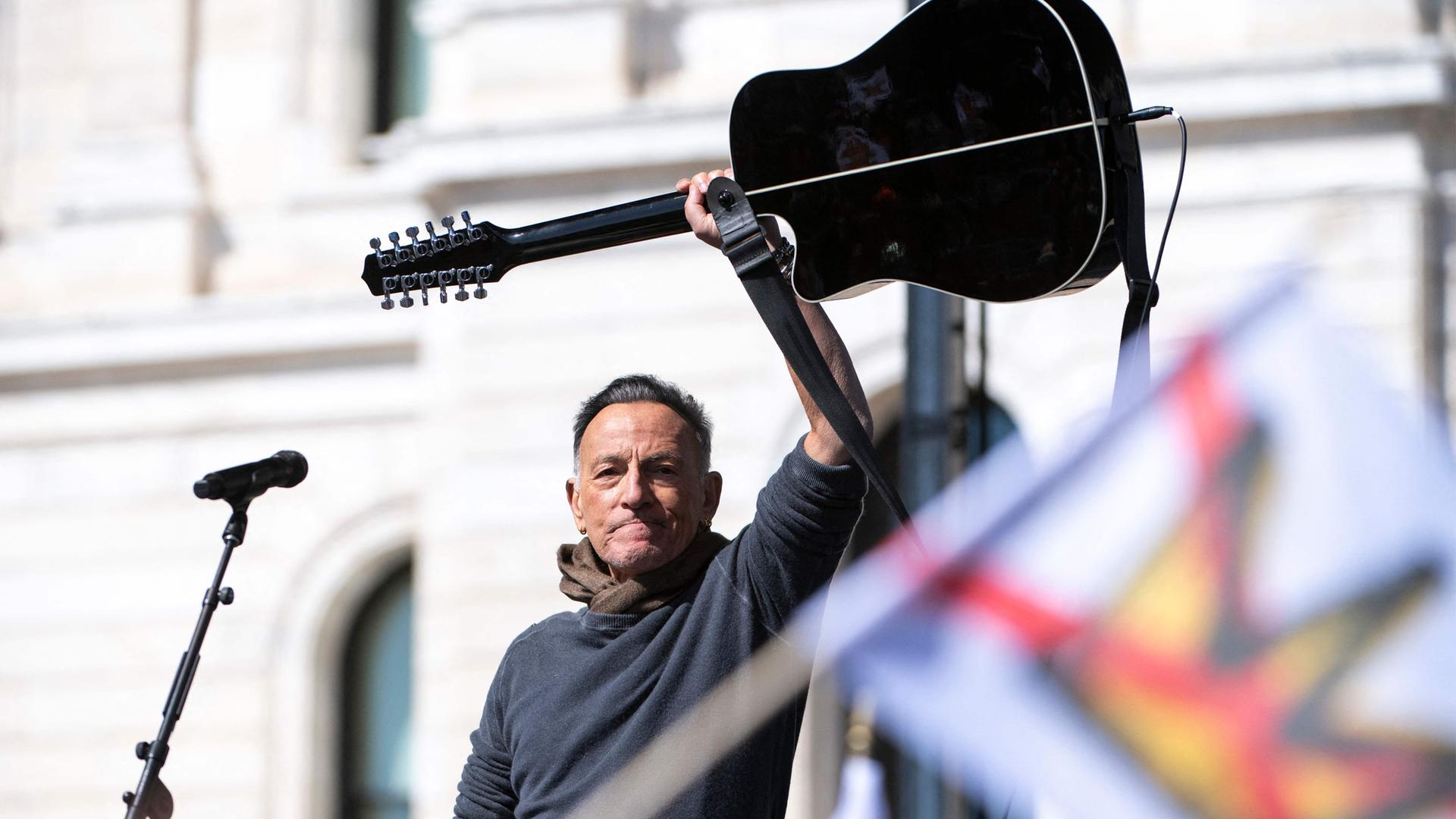 Mann hält Gitarre über den Kopf, im Vordergrund Fahne mit durchgestrichener Krone.