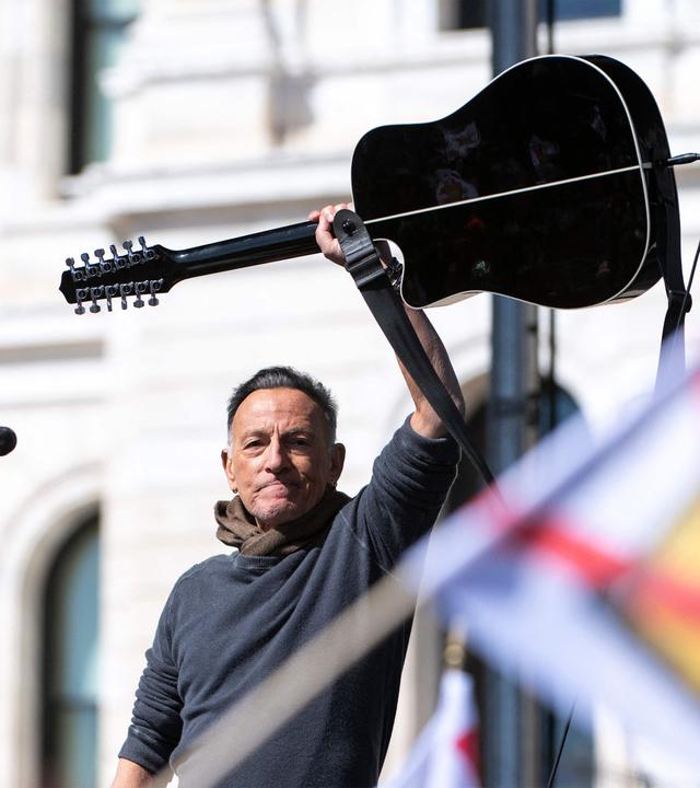 Mann hält Gitarre über den Kopf, im Vordergrund Fahne mit durchgestrichener Krone.